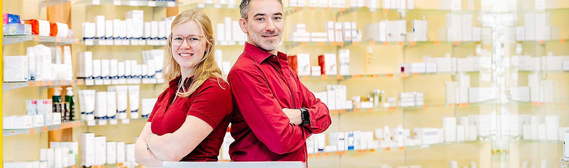 Zwei Mitarbeiter Rücken an Rücken mit verschränkten Armen im Verkaufsbereich der Apotheke